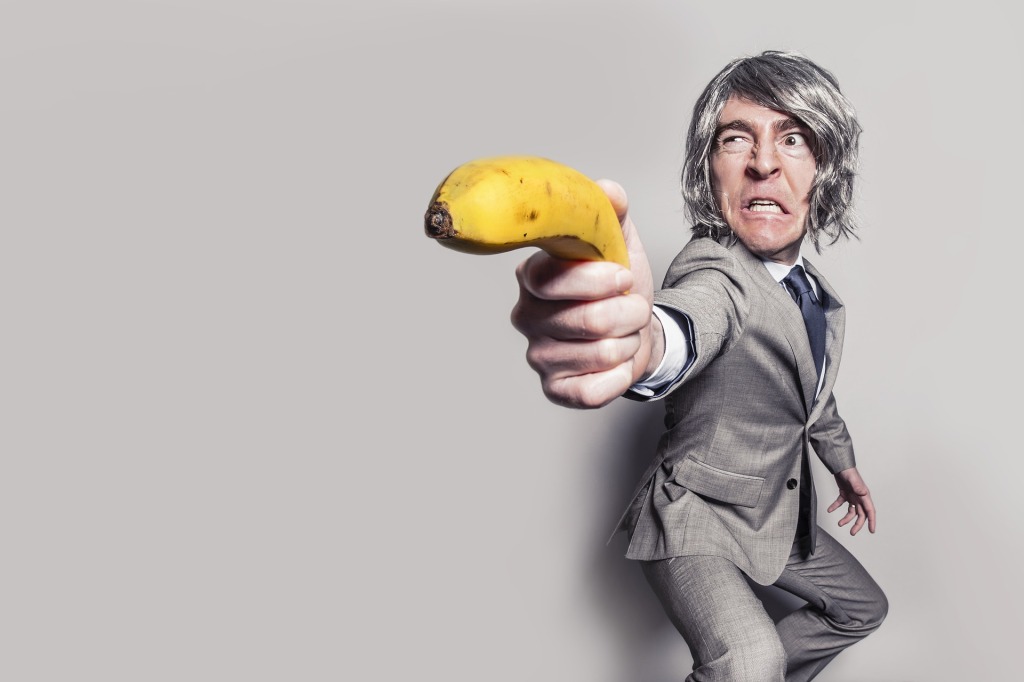 Gray haired man in a gray suit holding a banana as if it was a gun and making a "grr" face.