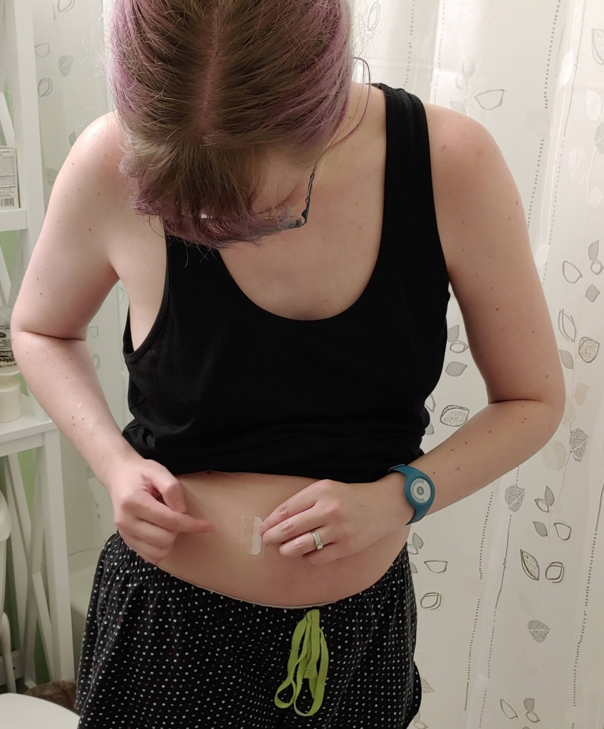 A white woman wearing a black tank top and pajama shorts, applying a translucent sticker to her stomach.